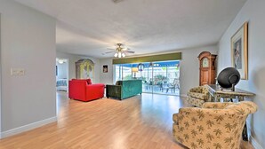 Un salon spacieux doté d’un parquet, d’un canapé rouge, d’un fauteuil vert et d’un autre fauteuil à motifs. On peut également apercevoir un ventilateur de plafond et une grande fenêtre offrant une belle vue.