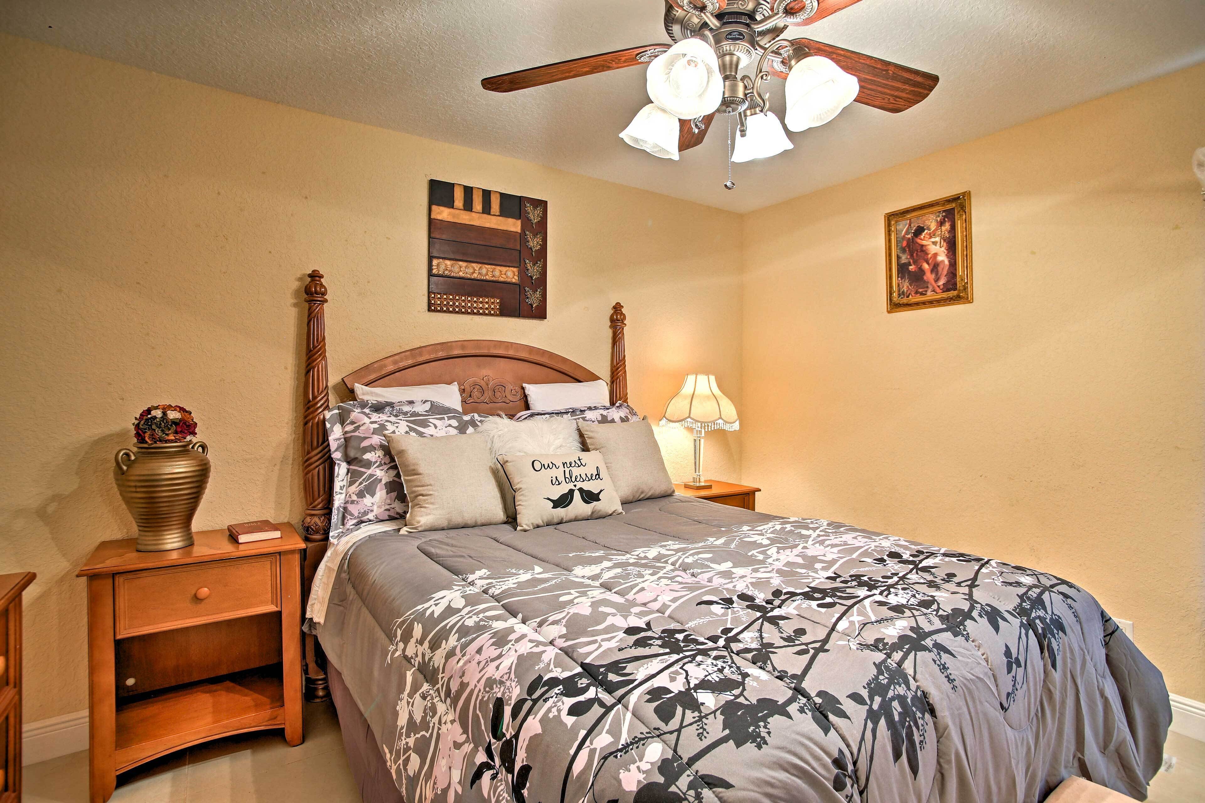 A bedroom with a wooden bed, a nightstand with a vase, a ceiling fan, and a wall-mounted lamp.