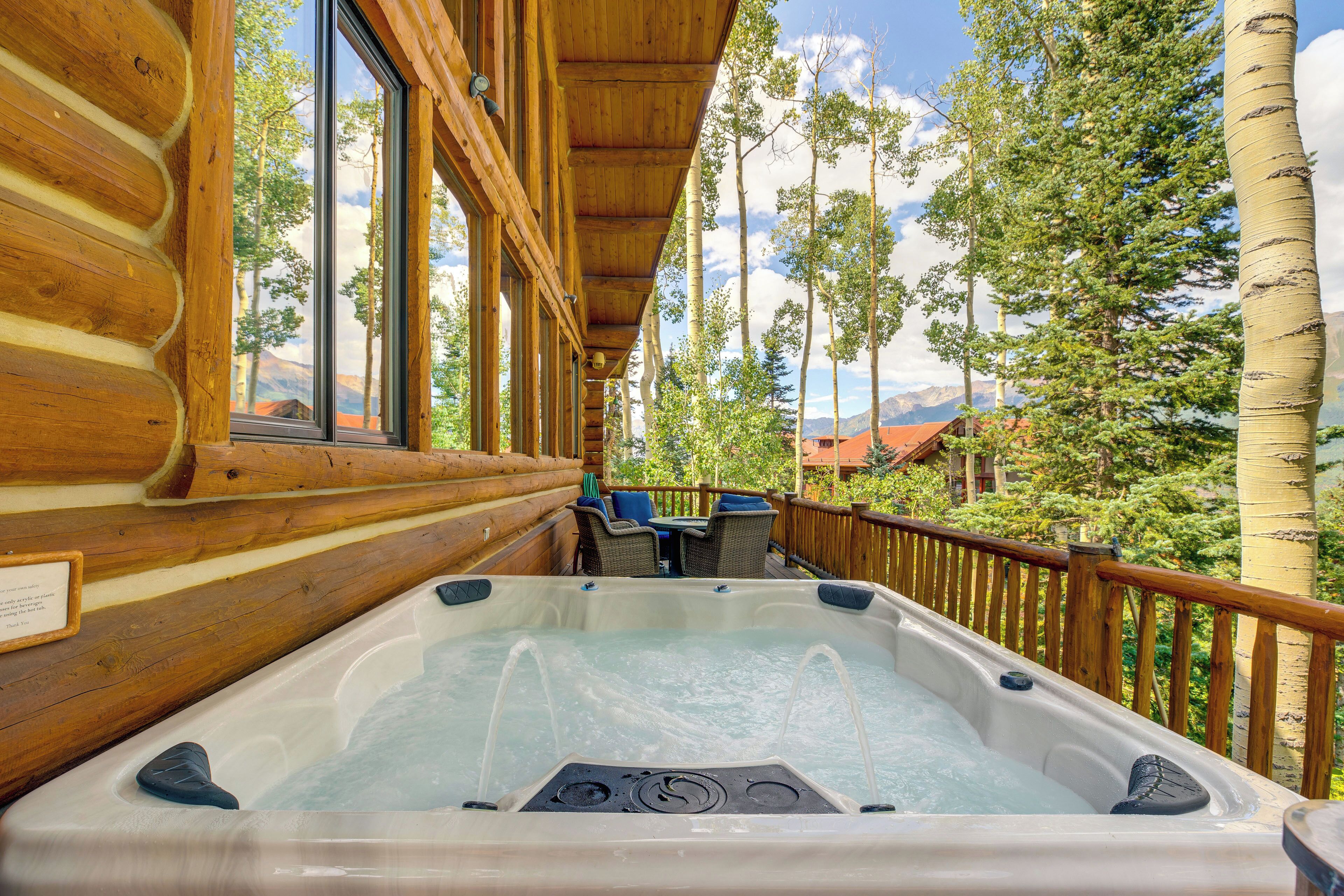 A wooden cabin with a hot tub on a deck, overlooking a mountainous landscape.