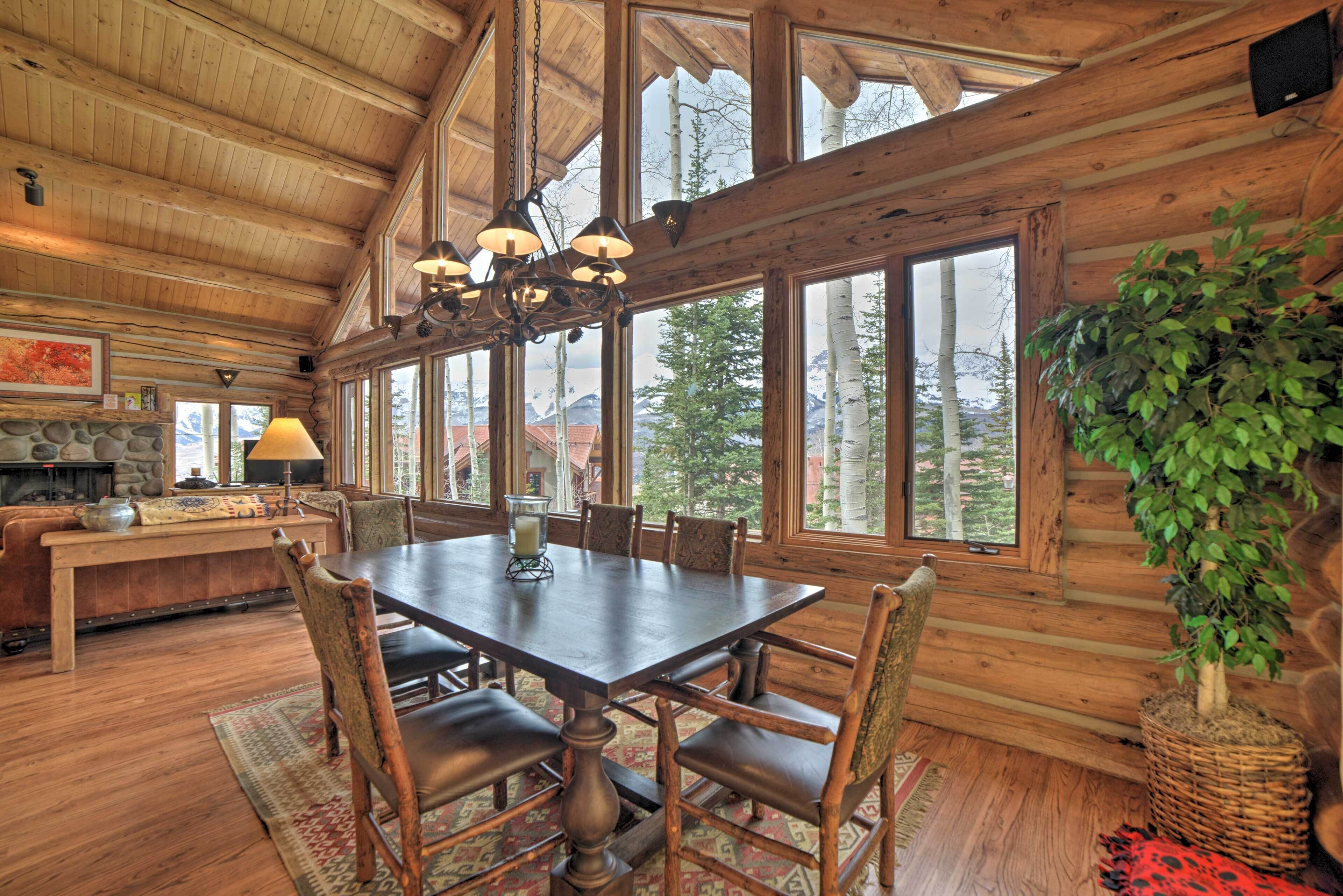 A log cabin interior with a dining table, chairs, a fireplace, and large windows offering mountain views.