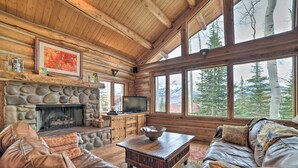 A log cabin interior with a stone fireplace, leather furniture, and large windows offering a mountain view. - Ski-in/ski-out Telluride Home w/ Deck & Hot Tub! (Telluride)