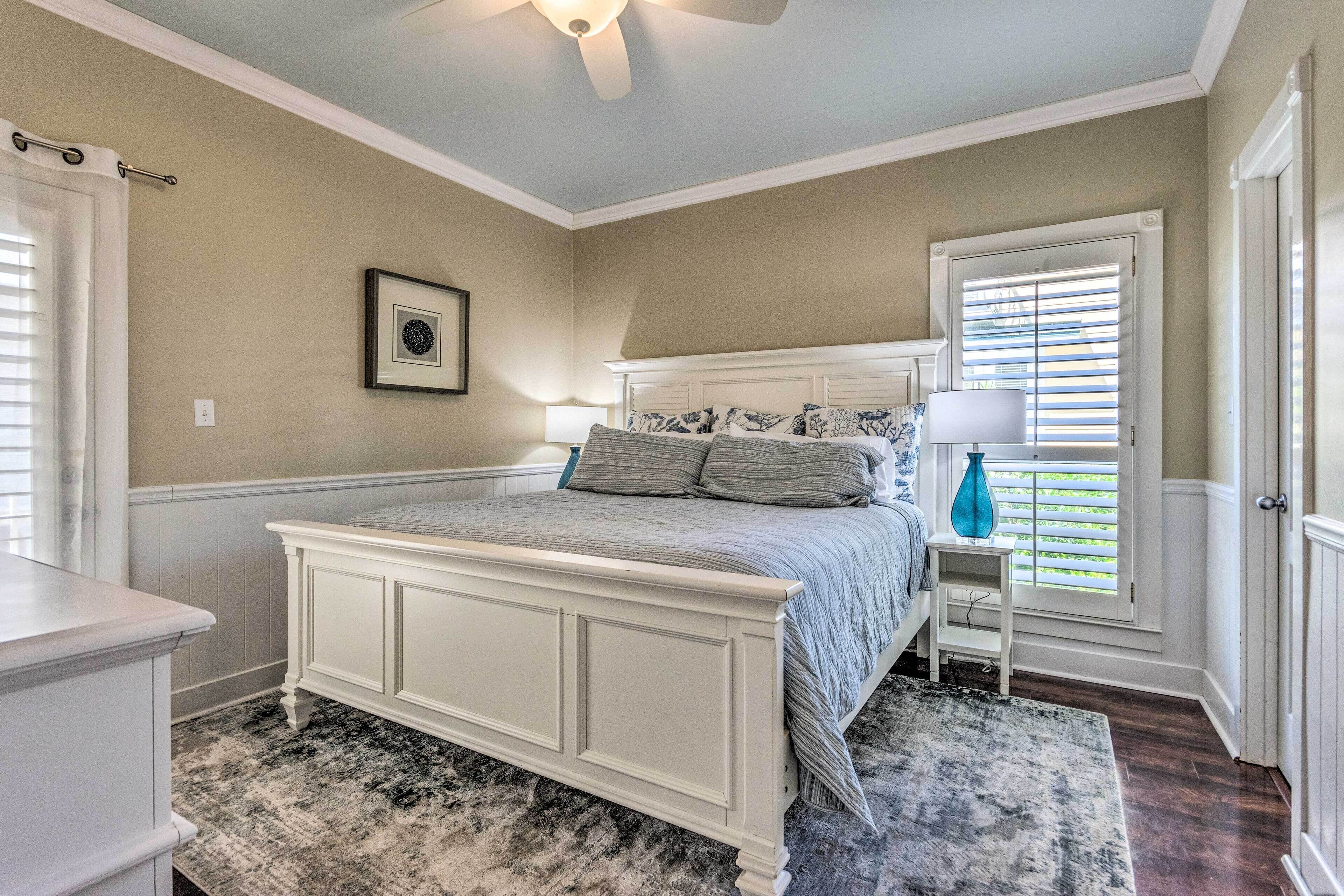 A bedroom with a white bed, a nightstand, a window with blinds, a ceiling fan, and a framed picture on the wall.