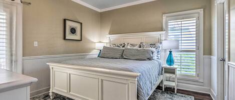A bedroom with a white bed, a nightstand, a window with blinds, a ceiling fan, and a framed picture on the wall.