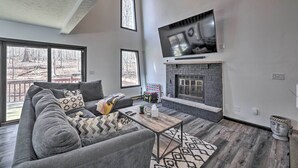 Una sala de estar moderna con un sofá gris, una chimenea, un televisor montado arriba y ventanas grandes que ofrecen vistas a árboles.