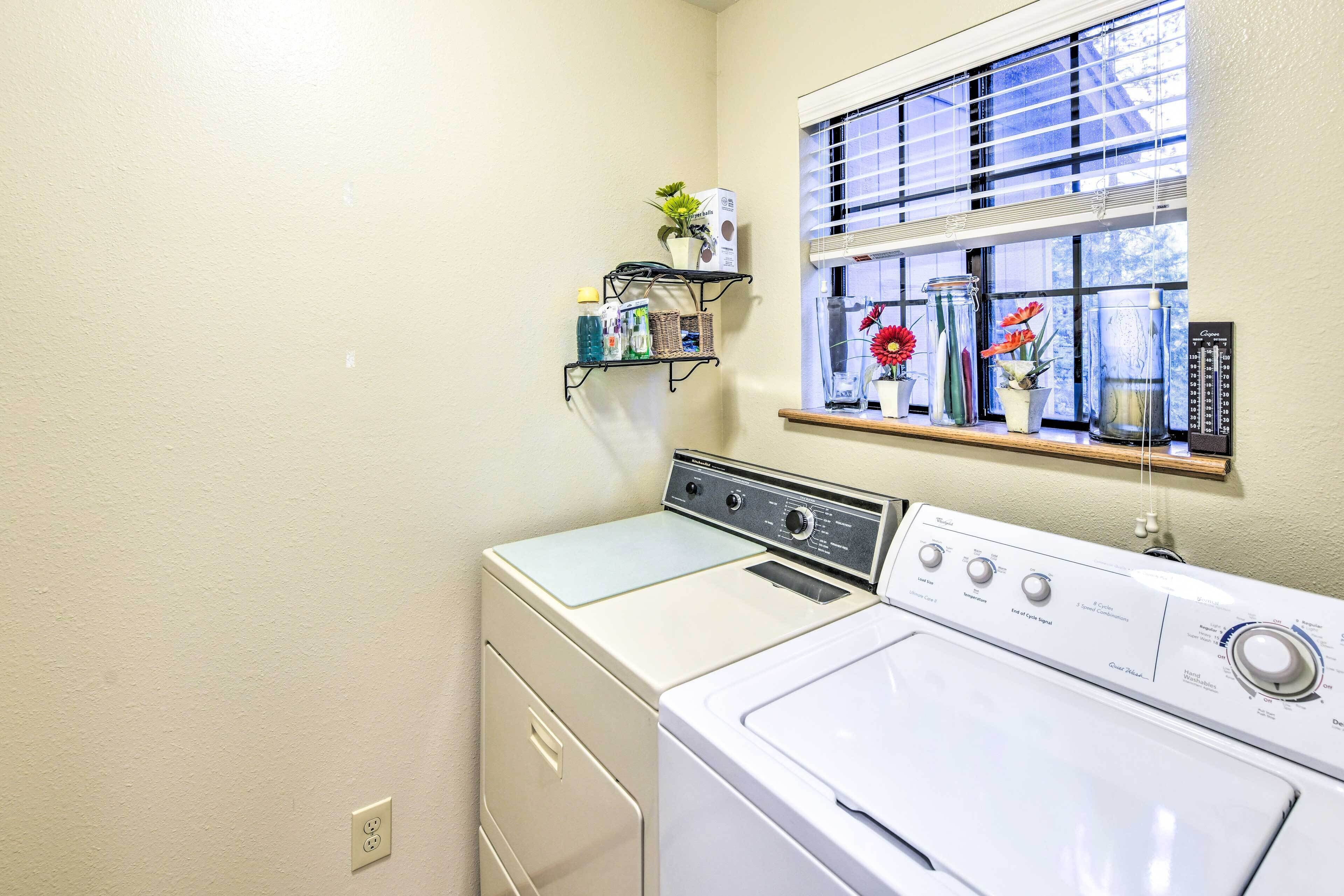 A laundry room with a front-loading washer and dryer, a window with blinds, and a shelf with various items.