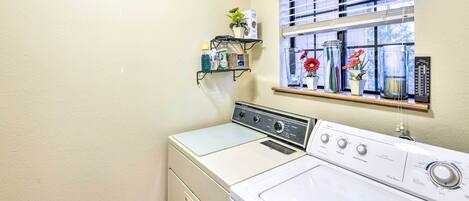 A laundry room with a front-loading washer and dryer, a window with blinds, and a shelf with various items.
