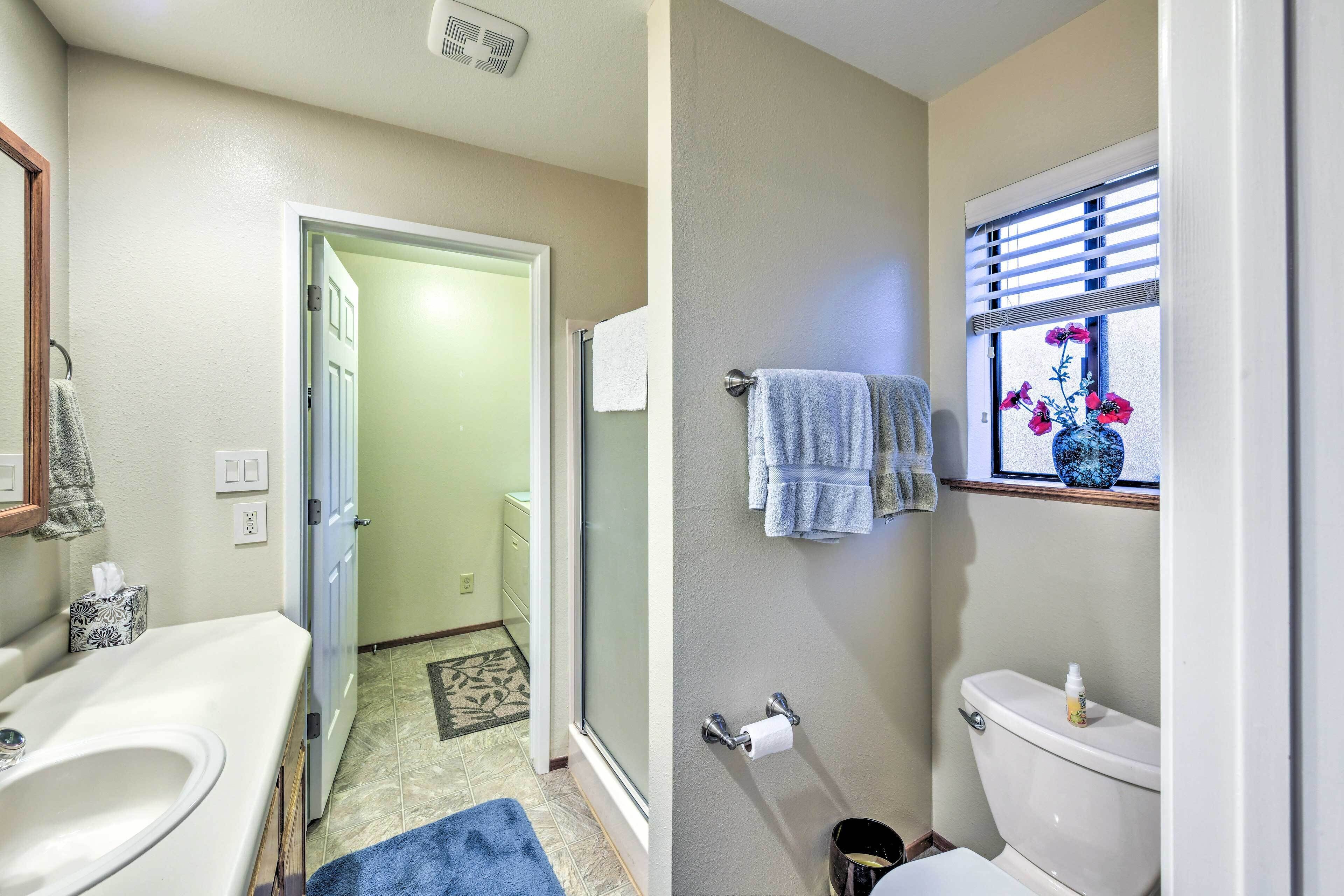 A bathroom with a white sink, toilet, and a window with blinds. There is a towel rack with towels and a small vase with flowers on the window sill.
