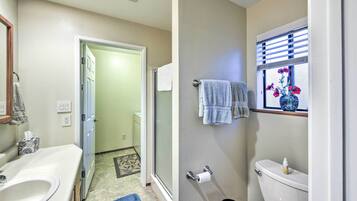 A bathroom with a white sink, toilet, and a window with blinds. There is a towel rack with towels and a small vase with flowers on the window sill.