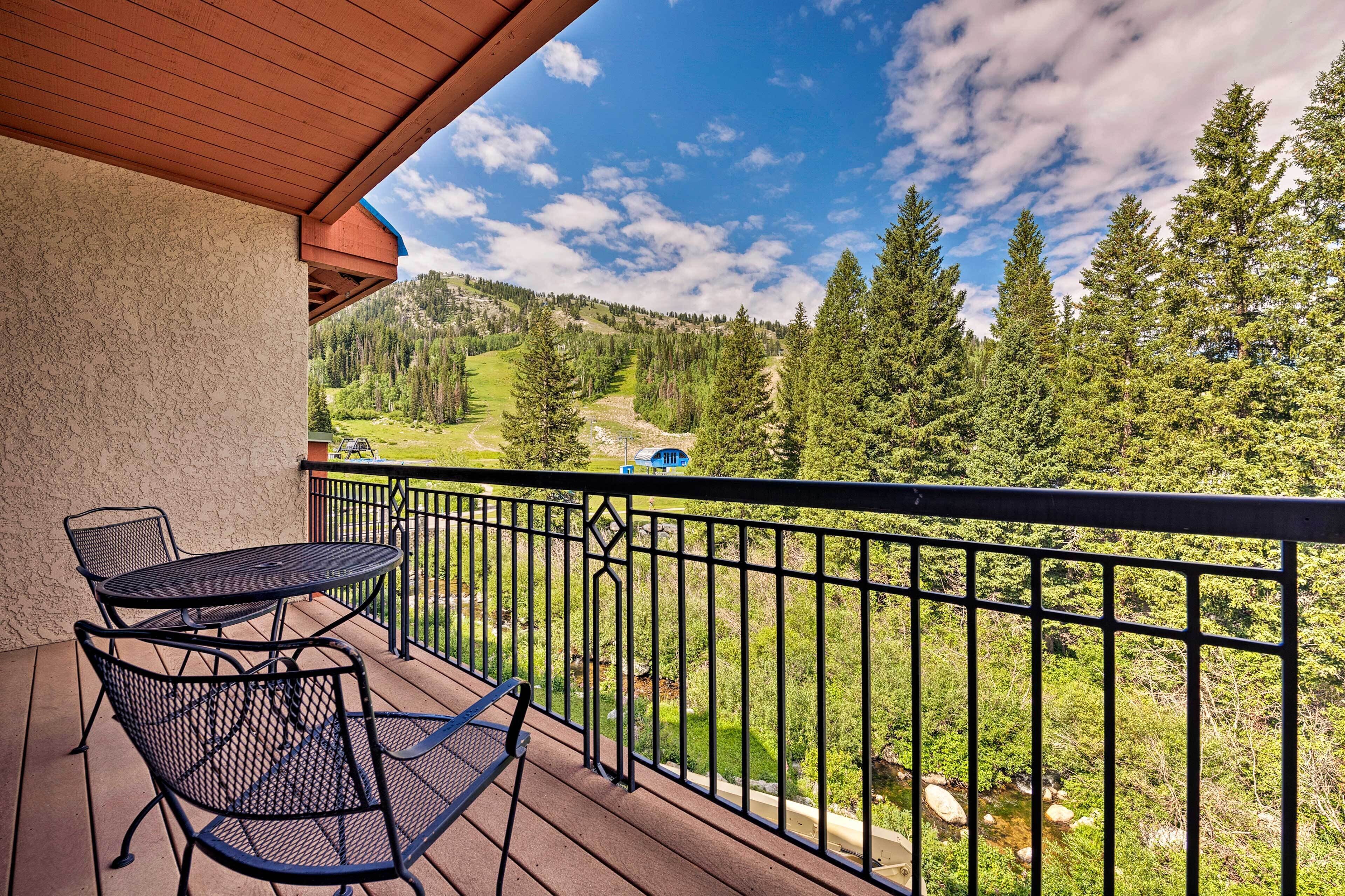 A balcony with a table and chairs, overlooking a mountainous landscape with trees and a clear sky.