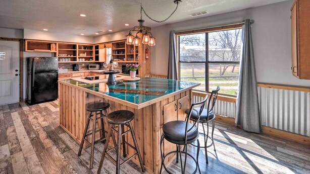 A kitchen with a central island, bar stools, wooden cabinets, and a large window with a view. - 'the Barn' Texas Wine Country Vacation Rental (Fredericksburg)