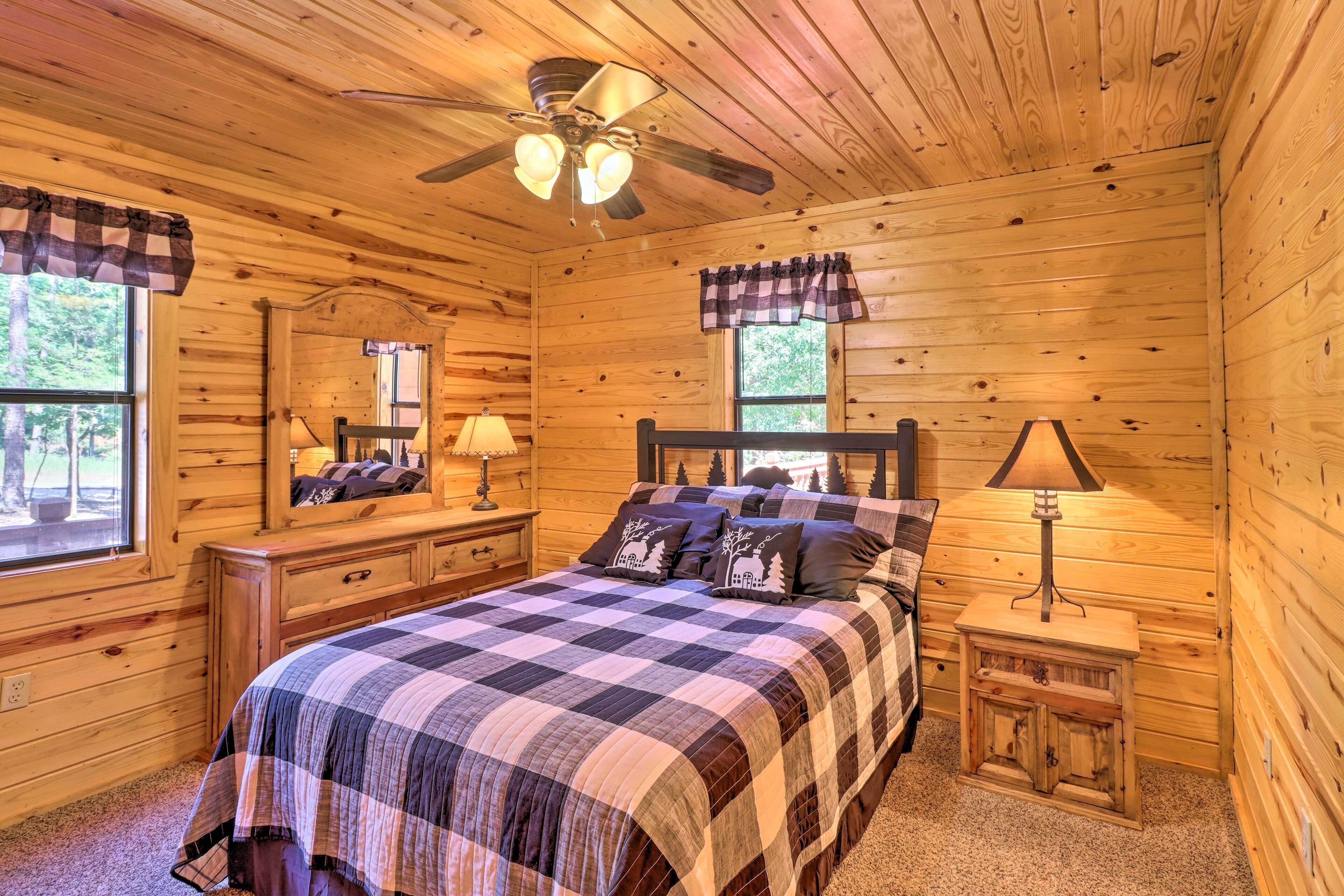A wooden cabin bedroom with a bed, two bedside tables, a ceiling fan, and a window with curtains.