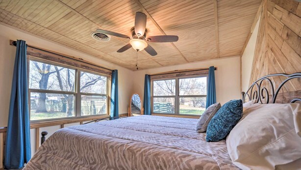 Une chambre dotée d’un ventilateur de plafond, de grandes fenêtres avec des rideaux, d’un lit avec une literie à motifs et d’une vue sur l’extérieur.