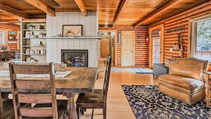 A rustic cabin interior with a wooden dining table, chairs, a fireplace, and a leather armchair.