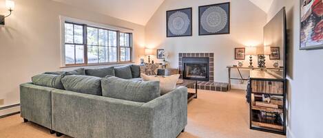 A spacious living room with a large grey sofa, a fireplace, and a bookshelf.