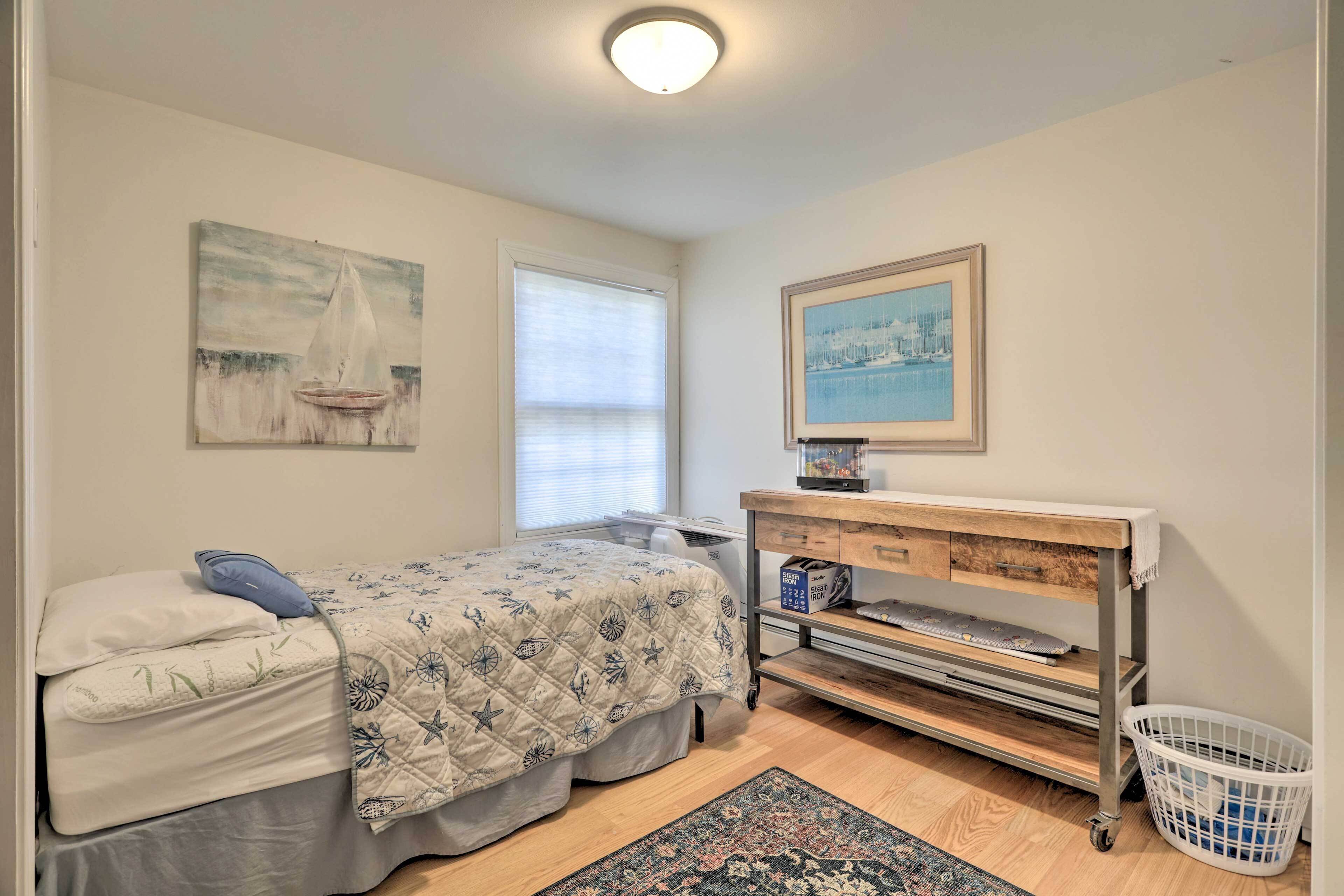 A bedroom with a bed, a nightstand, a desk, a chair, a window, and a wall-mounted picture.