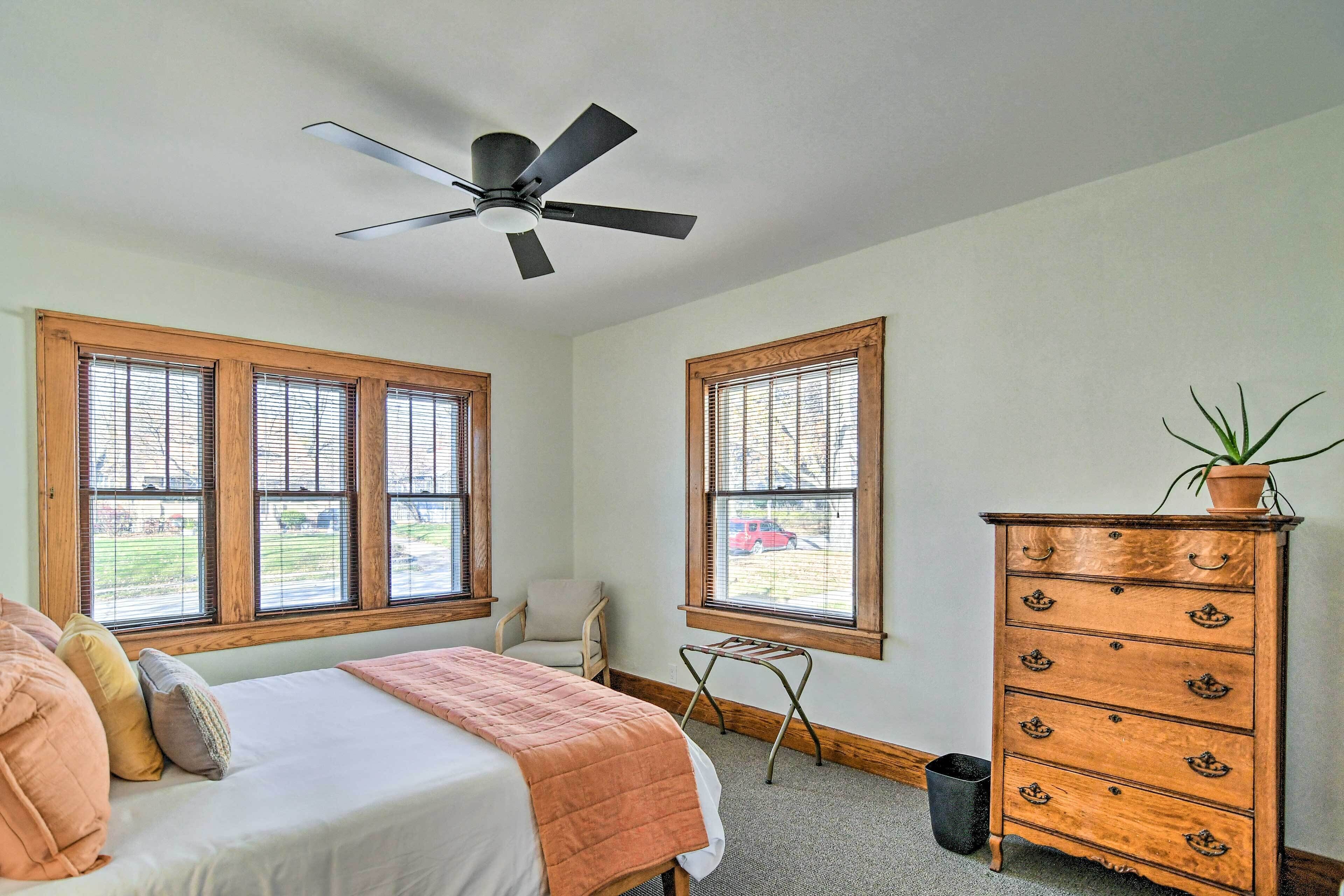 A bedroom with a bed, dresser, ceiling fan, windows, and a chair.