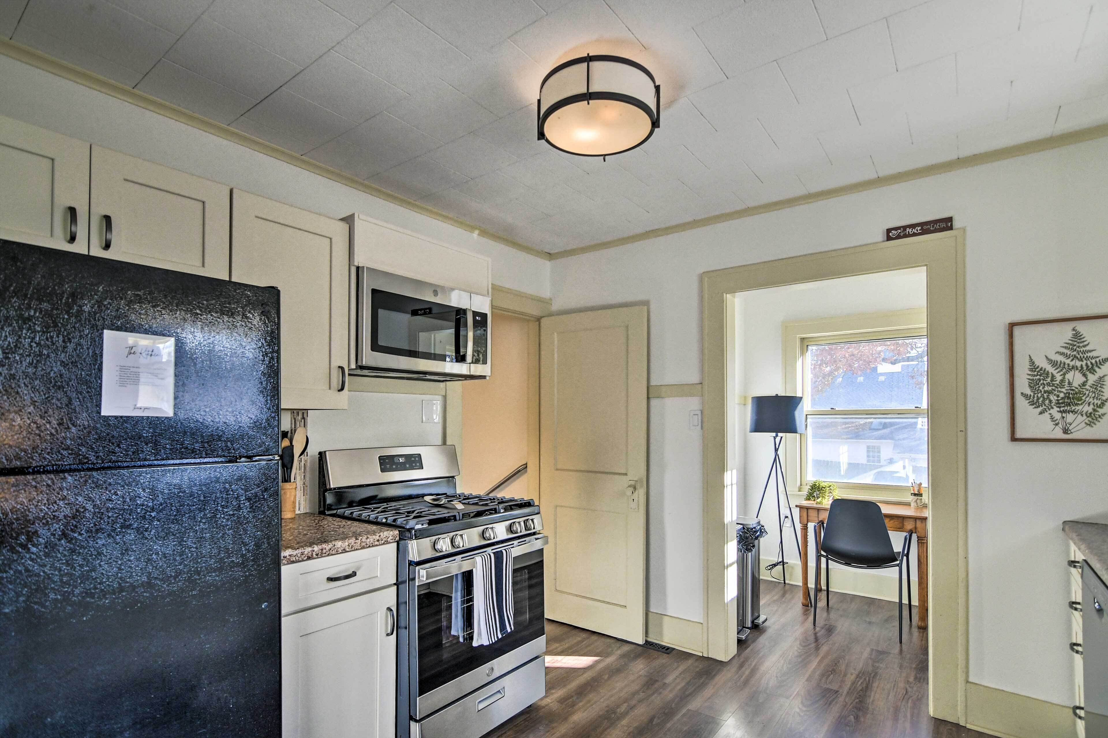 A kitchen with a refrigerator, microwave, stove, and oven. There is a door leading to another room, a desk with a chair, and a lamp.