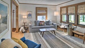 Un salon comprenant un canapé, des fauteuils, une table basse et un tapis. Il y a une fenêtre avec des stores, une lampe et un tableau encadré au mur.