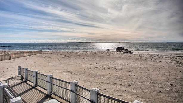 A beach with a wooden boardwalk and a rocky outcrop in the distance. - Rhode Island Oceanview Retreat: Walk to Shore! (Narragansett)