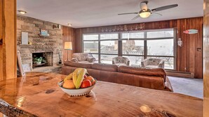 A cozy living room with a stone fireplace, wooden panel walls, a large window with a view, and a bowl of fruit on the counter. - Rhode Island Oceanview Retreat: Walk to Shore! (Narragansett)