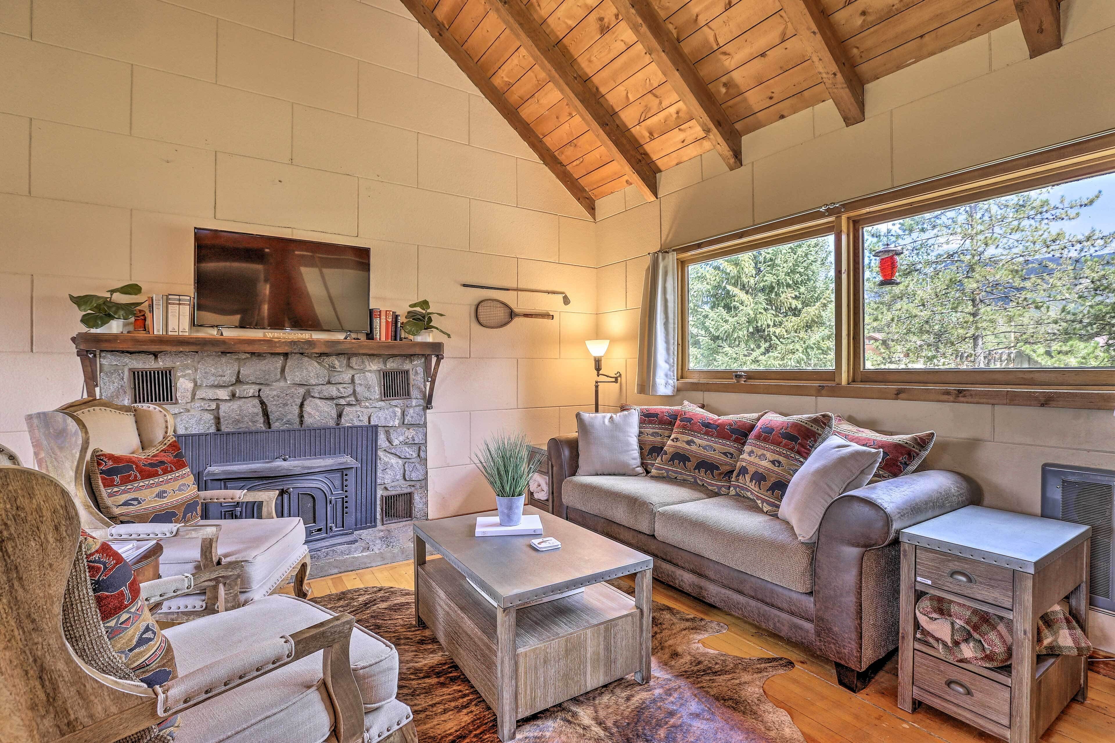 A cozy living room with a stone fireplace, wooden furniture, and a large window with a view of trees.
