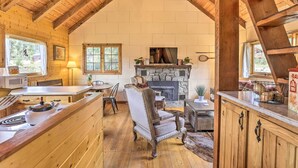 A cozy cabin interior with a stone fireplace, wooden beams, a dining area, and a kitchen with a stove. - Walk to Grand Lake: Historic Dtwn Cabin! (Grand Lake)