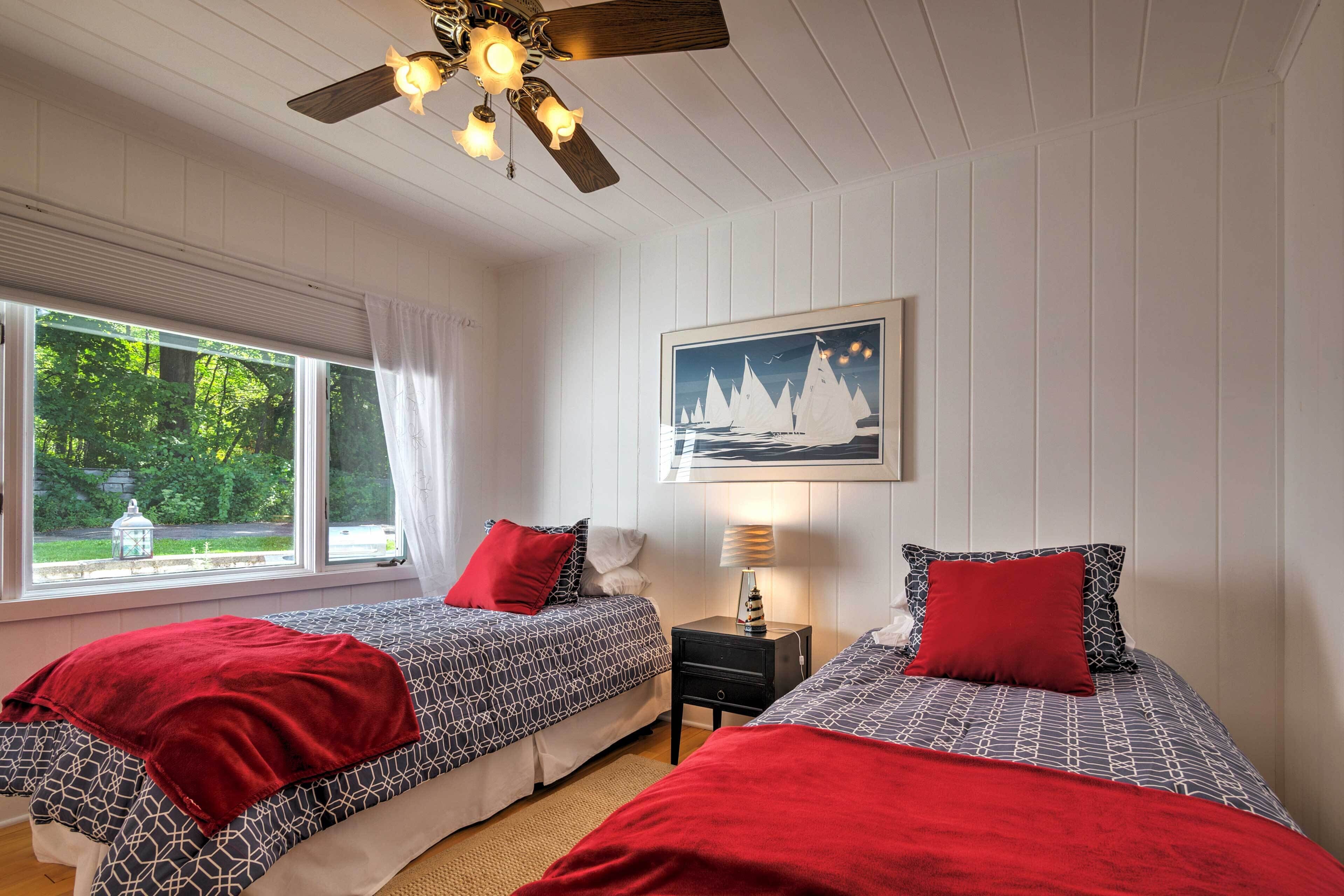 A bedroom with two beds, a ceiling fan, a window with a view of greenery, and a painting on the wall.