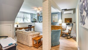 A cozy living room with a sofa, a rocking chair, and a wooden coffee table. There is a kitchen area with a refrigerator and shelves. A window with curtains is visible, and a ceiling fan is installed. - Bright Narragansett Beach Escape w/ Deck & Yard! (Narragansett)