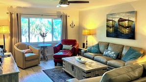 A living room with a sofa, armchairs, a coffee table, and a TV. There is a window with curtains, a painting on the wall, and a lamp. - Siesta Key Condo w/ Heated Pool < 1 Mi to Beach (Siesta Key)