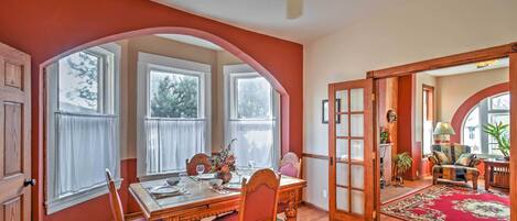 A dining area with a wooden table, chairs, and a ceiling fan. There are large windows with curtains, a red rug, and a view of the outdoors.