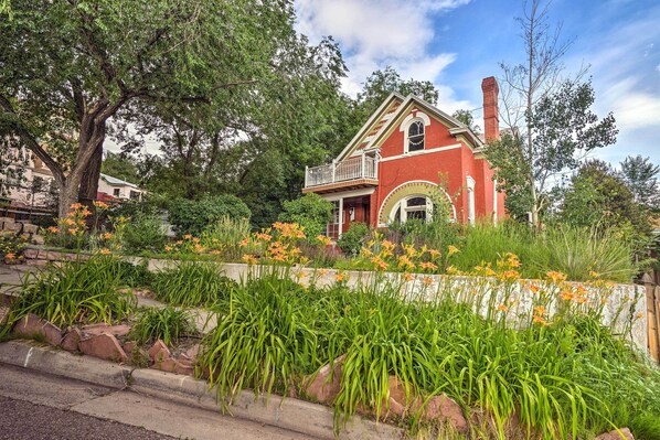 A red brick house with a balcony, surrounded by lush greenery and flowering plants.