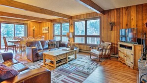 A cozy living room with wooden walls, large windows, a leather sofa, a wooden coffee table, and a TV stand. - 'peaceful Pines' Estes Park Home: Longs Peak View (Estes Park)