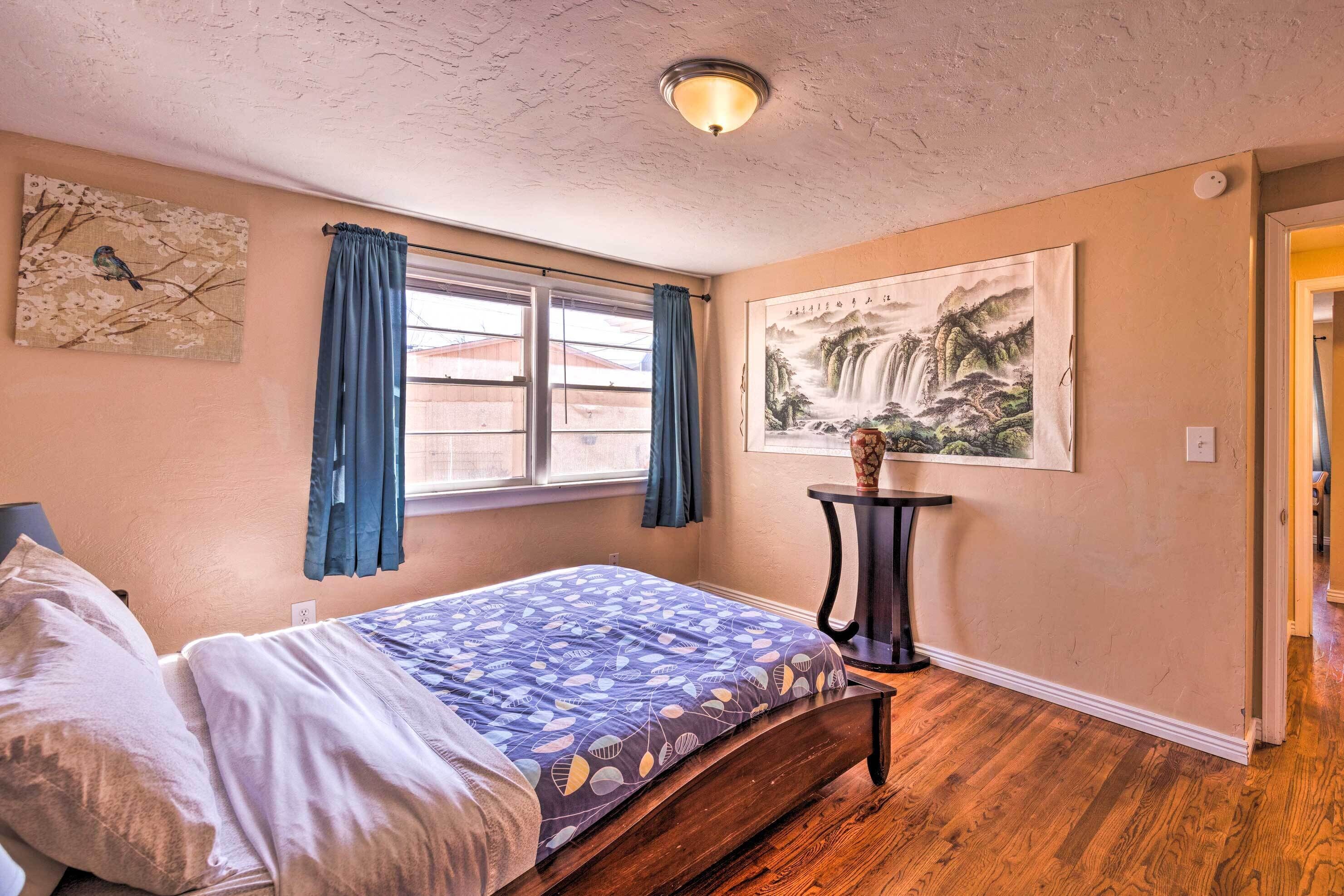 A bedroom with a bed, a window with curtains, a painting on the wall, and a small table with a vase.