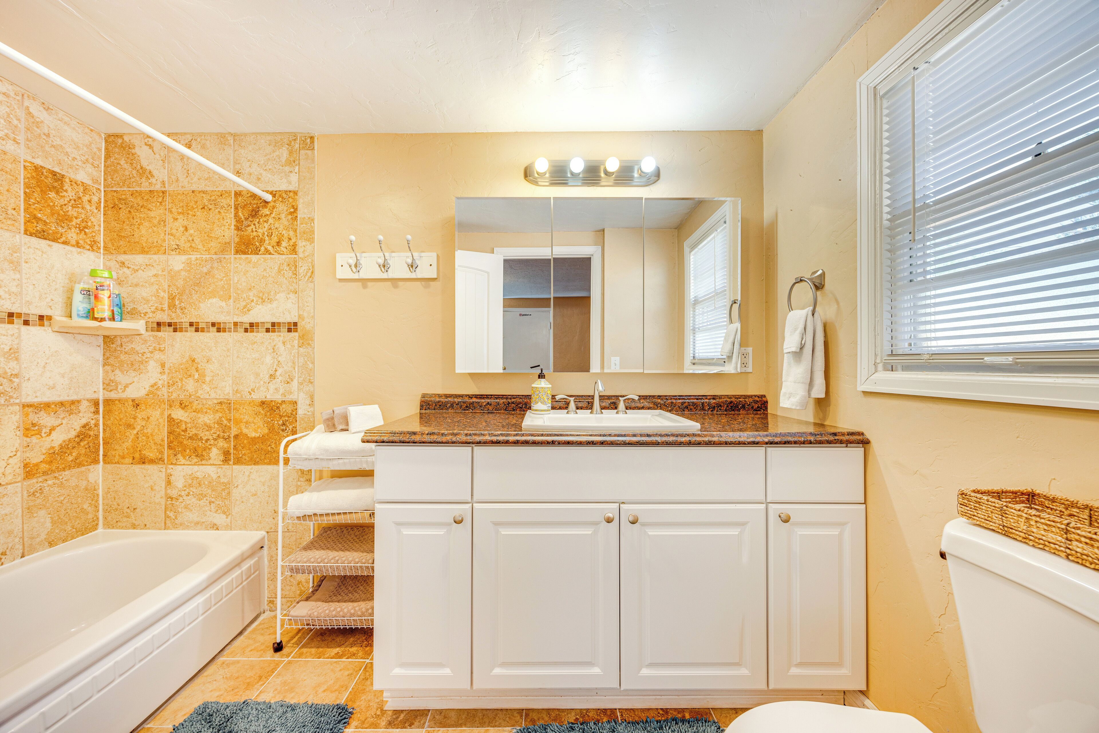 A bathroom with a bathtub, a sink with a mirror above it, and a toilet. There are shelves with toiletries and a window with blinds.