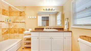 A bathroom with a bathtub, a sink with a mirror above it, and a toilet. There are shelves with toiletries and a window with blinds.