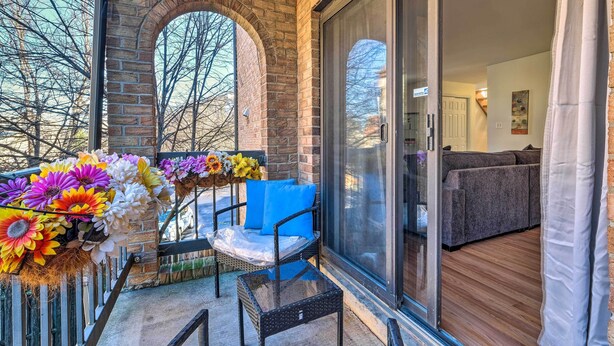A balcony with a brick wall, a glass door leading to a living area, and a table with a chair and a vase of flowers.