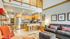 A spacious living area with a grey sofa, ottoman, and a red 'Gone Skiing' throw pillow. A kitchen with wooden cabinets and stainless steel appliances is visible in the background. - Ski & Relax Fireside: Frisco Gem w/ Resort Perks! (Frisco)