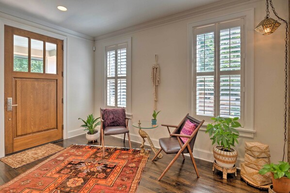 A spacious room with a wooden door, large windows with blinds, a patterned rug, two chairs, and potted plants.