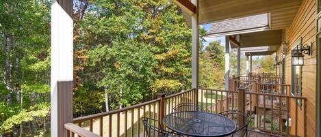 A wooden deck with a round table and chairs, surrounded by a wooden railing, overlooking a forested area.
