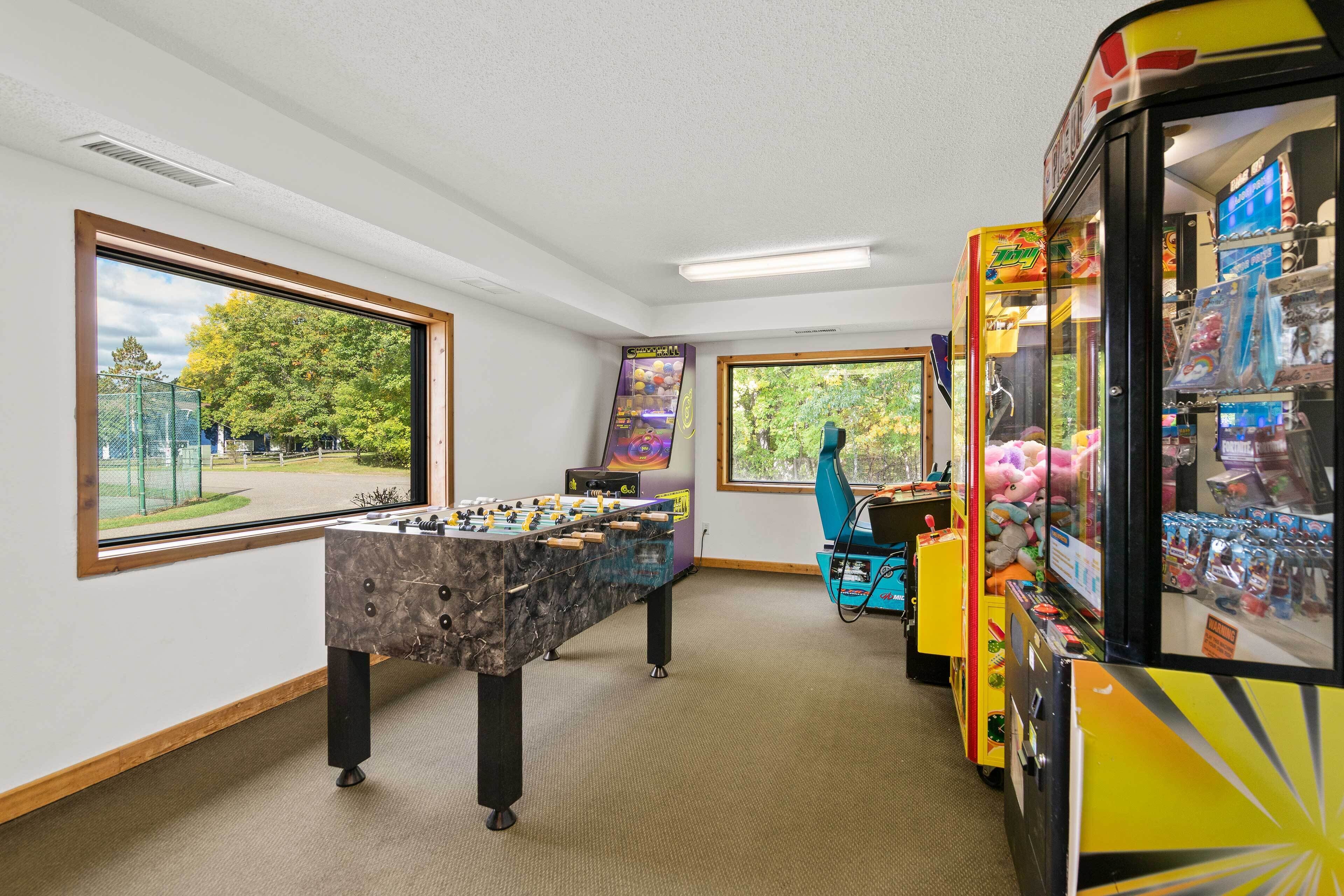 A room with a foosball table, vending machines, and large windows offering views of a playground and trees.