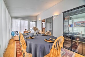 A dining area with a round table set for six, wooden chairs, a glass cabinet with board games, and a view through the window.