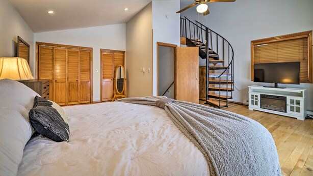 A bedroom with a bed, a ceiling fan, a TV, wooden closets, a staircase, and a window with blinds.