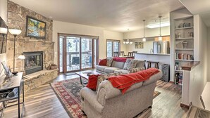 A spacious living room with a stone fireplace, a large sofa, and a dining area with a kitchen in the background. - The Chelsea Condo w/ Hot Tub & Pool Access! (Breckenridge)