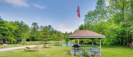A gazebo with a flagpole, picnic tables, and a pond in a park.