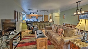 A living room with a sofa, ottoman, and a TV on a stand. There is a kitchen in the background with a refrigerator and a dining area. - < 2 Mi to Frisco Adventure Park: Cozy Townhome! (Frisco)
