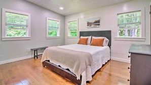 Une chambre à coucher avec un grand lit, un banc et une commode. Il y a trois fenêtres avec des stores et un tableau au mur.