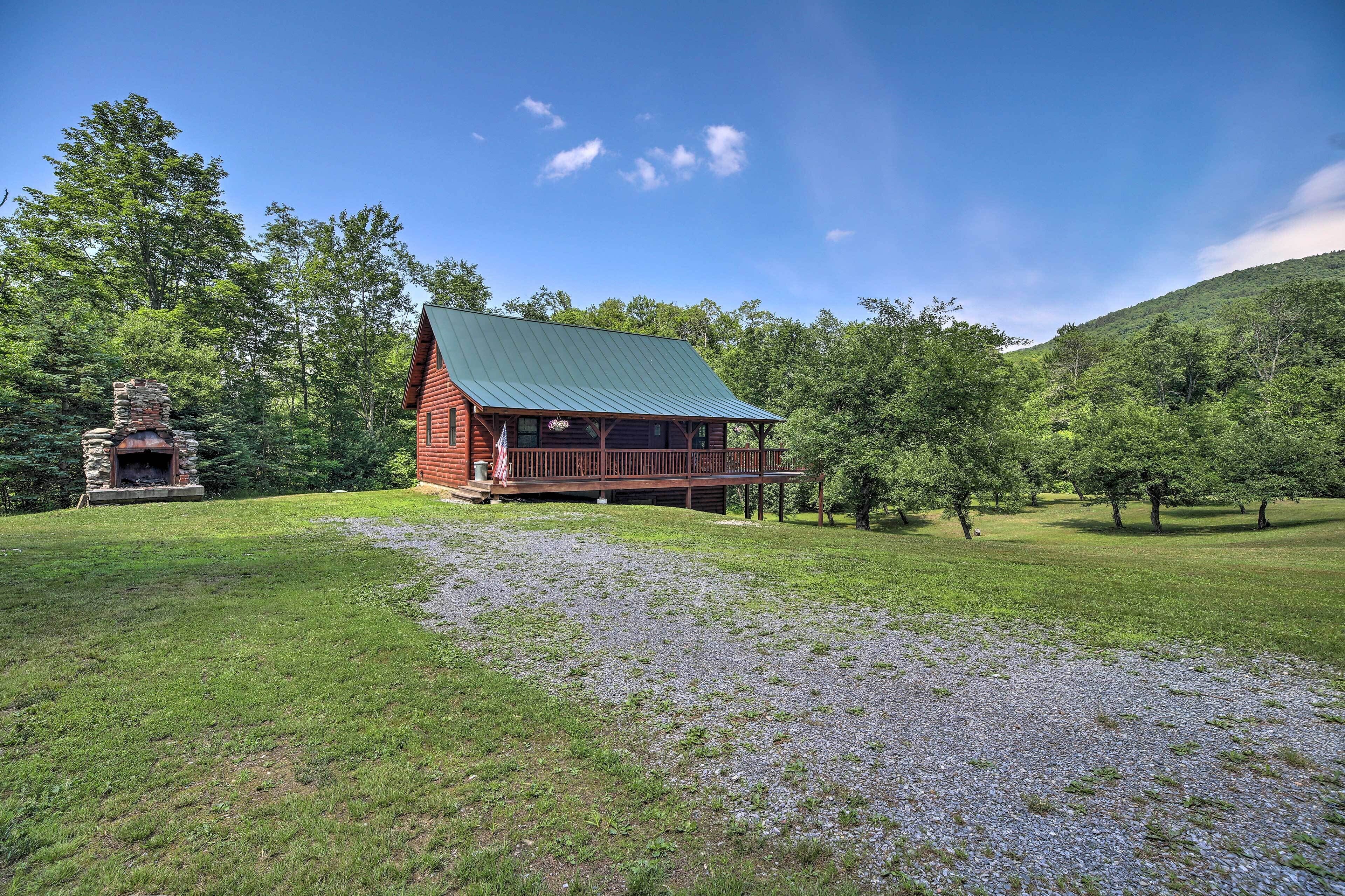 A log cabin with a stone fireplace, surrounded by a grassy area and trees.