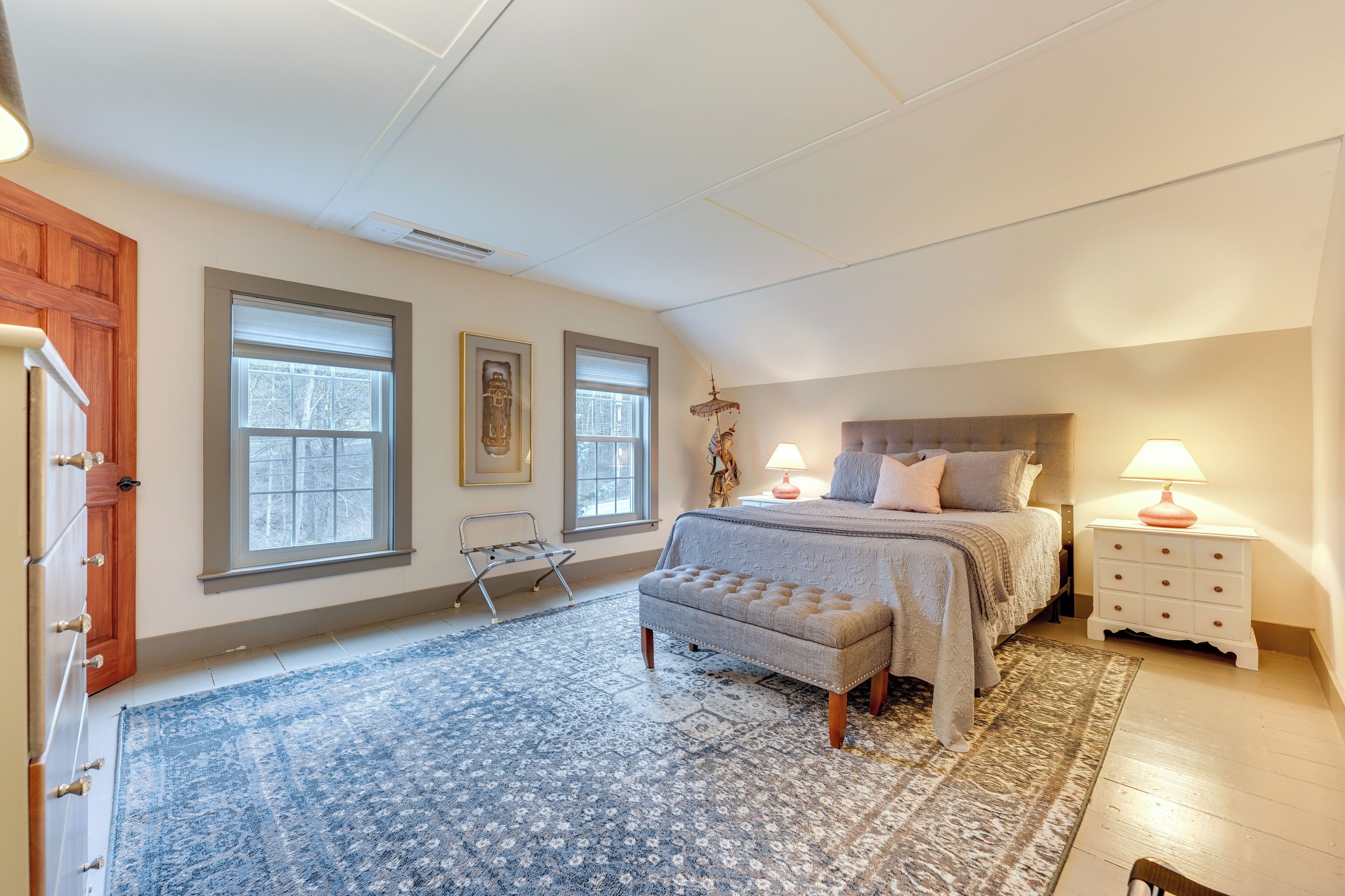 A bedroom with a bed, nightstand, lamp, bench, and two windows.