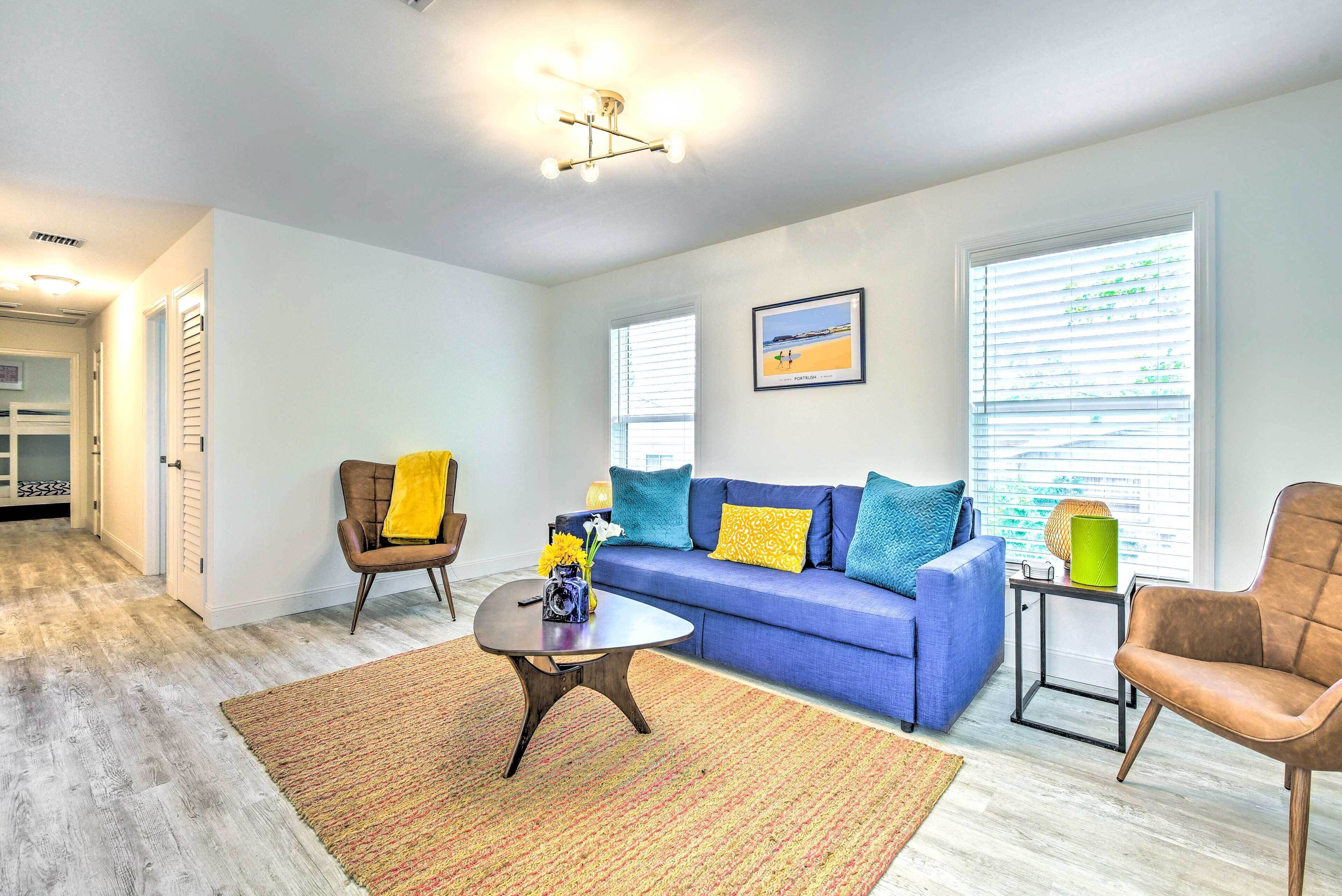 A living room with a blue sofa, a brown armchair, a coffee table, and a rug. There are two windows with blinds, a ceiling light fixture, and a framed picture on the wall.