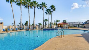 A swimming pool with clear blue water, surrounded by palm trees and blue lounge chairs. - Furnished Deck: Waterfront Port Isabel Cottage! (Port Isabel)
